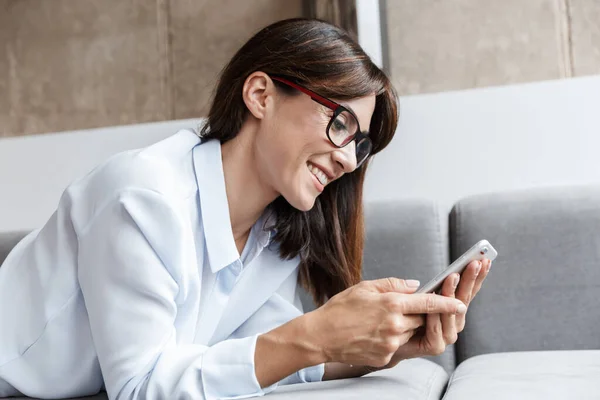 Alegre feliz mujer de negocios en el interior de casa se encuentra en el sofá en la sala de estar utilizando el teléfono móvil . —  Fotos de Stock