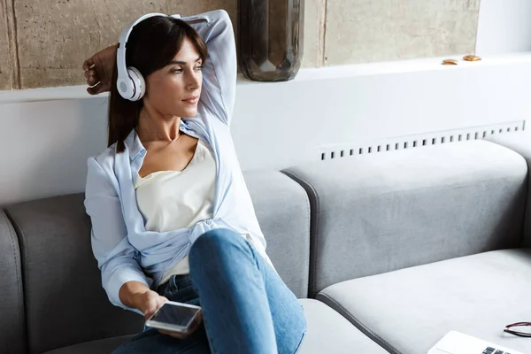 Mujer en el interior de casa sentarse en el sofá en la sala de estar escuchando música con auriculares utilizando el teléfono móvil . — Foto de Stock