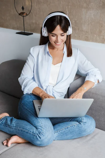 Donna in casa seduta sul divano in salotto ad ascoltare musica con le cuffie utilizzando il computer portatile — Foto Stock
