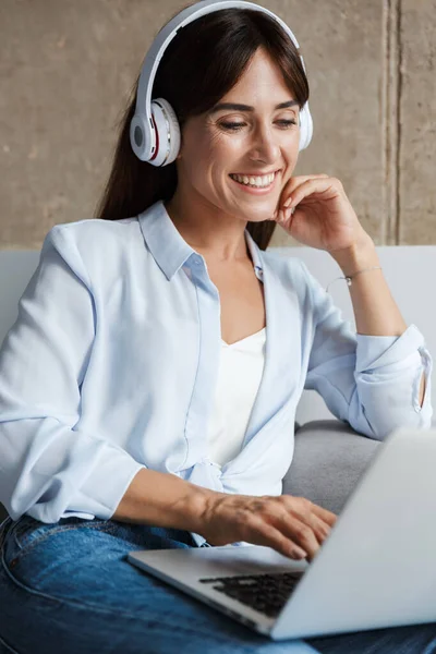 家里的女人坐在客厅的沙发上，用手提电脑用耳机听音乐 — 图库照片