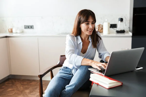 Mulher sorridente dentro de casa na cozinha usando o computador portátil . — Fotografia de Stock