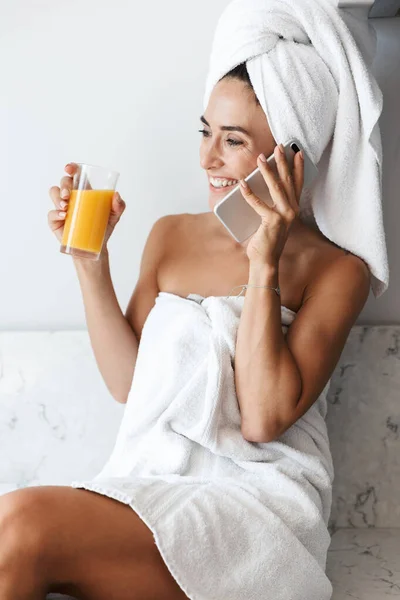 Happy woman in towel after spa procedure shower indoors at home at the kitchen drinking juice talking by mobile phone. — Stockfoto