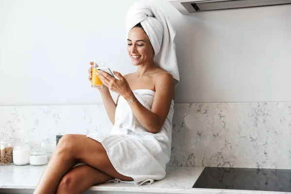 휴대폰을 사용 해 부엌에서 집에 있는 주스를 마시며 온천 목욕을 한 행복 한 여성 이 수건으로 웃고 있는 모습. — 스톡 사진