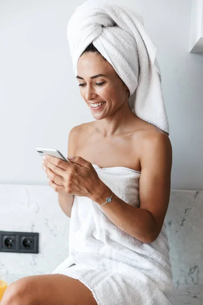 휴대폰을 사용하여 부엌에서 집안에서 샤워를 하고 난 뒤 행복 한 여인 이 수건으로 웃고 있는 모습. — 스톡 사진