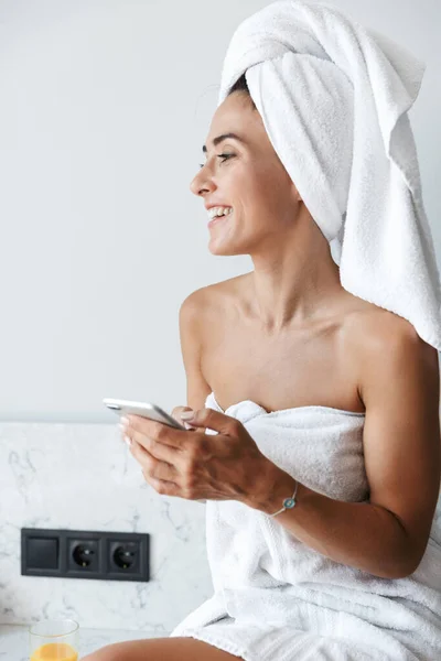 Χαμογελώντας ευτυχισμένη γυναίκα με πετσέτα μετά το ντους διαδικασία spa σε εσωτερικούς χώρους στο σπίτι στην κουζίνα χρησιμοποιώντας το κινητό τηλέφωνο. — Φωτογραφία Αρχείου