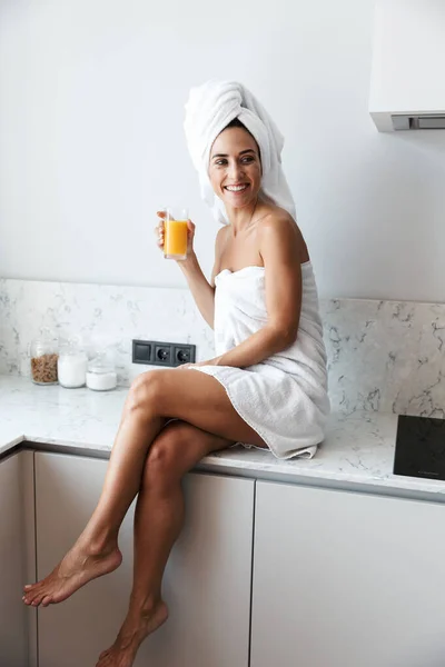Позитивная улыбающаяся женщина в полотенце после процедуры спа душ в помещении дома на кухне питьевой сок . — стоковое фото