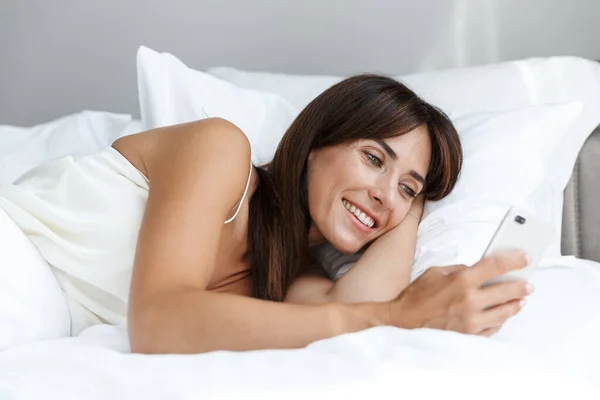 Šťastná veselá spokojená žena doma v posteli pomocí mobilního telefonu. — Stock fotografie