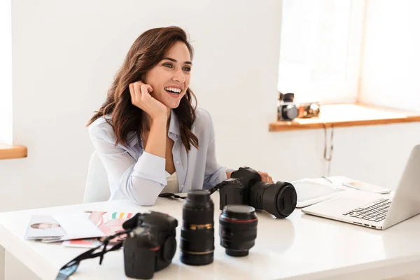 Photographer woman in office sit at the table near camera and lenses.