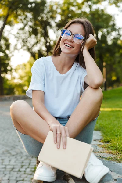 Usmívající se mladá dívka drží knihu, zatímco sedí v parku — Stock fotografie