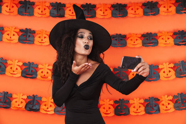 Image of cute witch girl in black halloween costume taking selfi