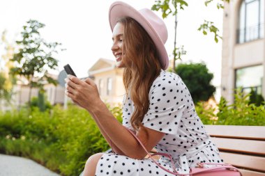 Kızıl saçlı kadın bankta oturup cep telefonuyla sohbet ediyor..