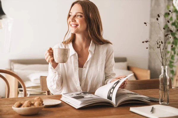 Frau sitzt drinnen im Büro und liest Zeitschrift und trinkt Kaffee. — Stockfoto