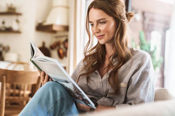 Positiv optimistische rothaarige Frau drinnen sitzt zu Hause und liest Buch oder Zeitschrift. — Stockfoto
