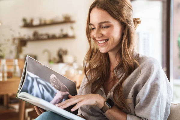 Positiv optimistische rothaarige Frau drinnen sitzt zu Hause und liest Buch oder Zeitschrift. — Stockfoto