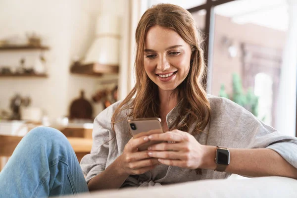Lachende vrolijke mooie roodharige vrouw thuis met behulp van mobiele telefoon. — Stockfoto