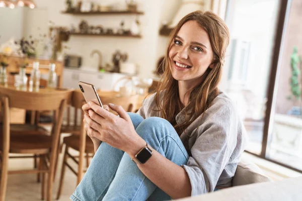 Sonriente alegre hermosa pelirroja en casa usando el teléfono móvil . — Foto de Stock