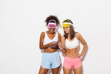 İki neşeli çokuluslu kadının portresi. Güneşlik şapkalar takıp cep telefonlarına yazıyorlar ve gülüyorlar.