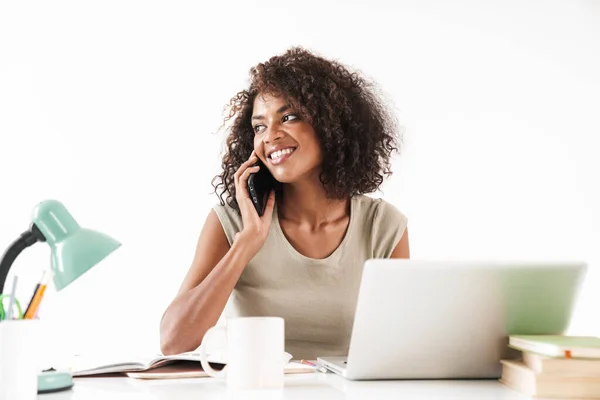 Sorrindo jovem mulher africana usando computador portátil enquanto toma uma xícara de café e sentado na mesa isolado sobre fundo branco e falando no telefone celular — Fotografia de Stock