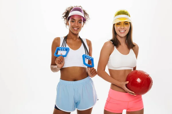 Портрет двух улыбающихся мультиэтнических женщин в козырьковых шляпах, держащих фитнес-мяч и экспандер — стоковое фото
