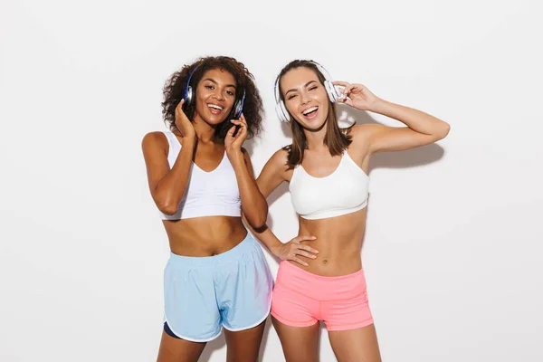 Портрет двух веселых мультиэтнических женщин в спортивной одежде, смеющихся и слушающих музыку в наушниках — стоковое фото