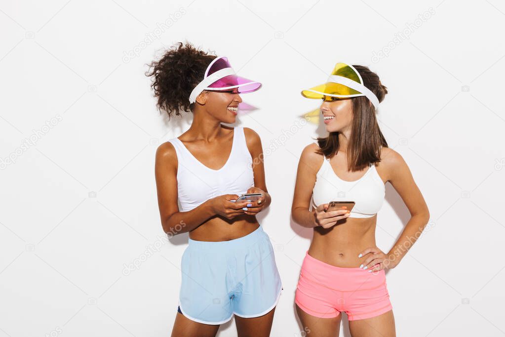 Portrait of two laughing multinational women wearing visor hats typing on cellphones and looking at each other