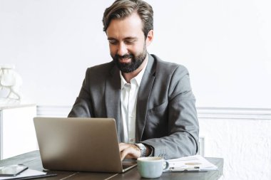Ofiste çalışırken laptopta yazan ve kahve içen neşeli, kendinden emin iş adamı görüntüsü