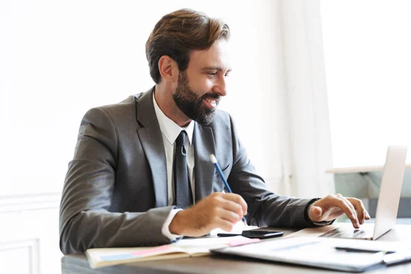 Image of smiling confident businessman working at laptop in office while writing down notes — Stock Photo, Image
