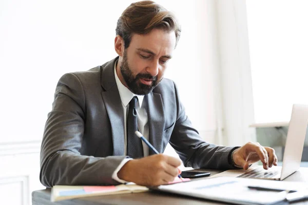 Image of serious confident businessman working at laptop in office while writing down notes — ストック写真