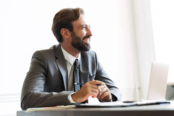 Image of laughing confident businessman looking aside while working at laptop in office — Stock Photo, Image
