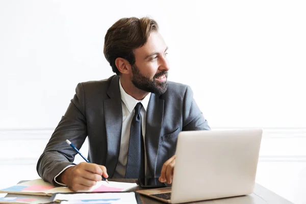 Beeld van lachende bebaarde zakenman die werkt op laptop in kantoor tijdens het opschrijven van notities — Stockfoto