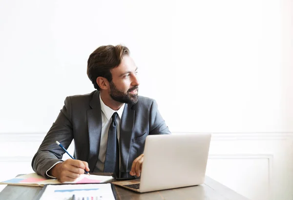 Image of satisfied bearded businessman working at laptop in office while writing down notes and looking aside — ストック写真