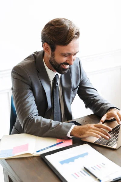 Przystojny młody pozytywny biznesmen siedzi w biurze za pomocą laptopa pracy w pomieszczeniach. — Zdjęcie stockowe