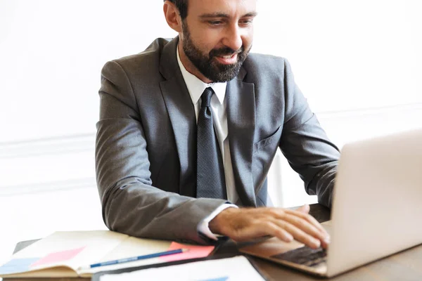 Beau jeune homme d'affaires positif assis au bureau en utilisant un ordinateur portable travaillant à l'intérieur . — Photo