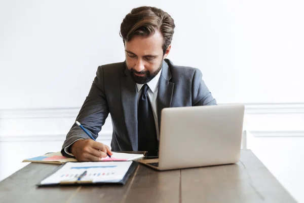 Beeld van geconcentreerde mannelijke zakenman die werkt op laptop en mobiele telefoon in kantoor tijdens het opschrijven van notities — Stockfoto