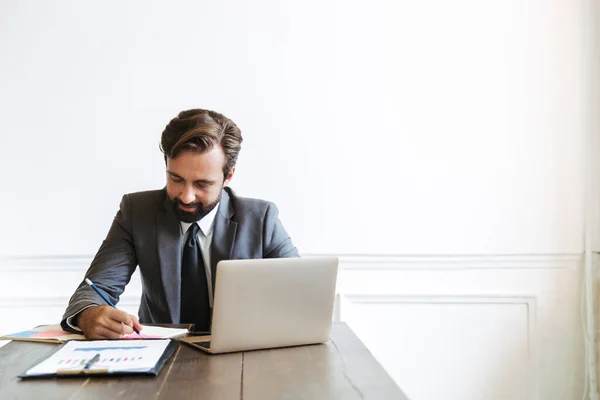 Image d'un homme d'affaires masculin non rasé travaillant à un ordinateur portable au bureau tout en écrivant des notes — Photo