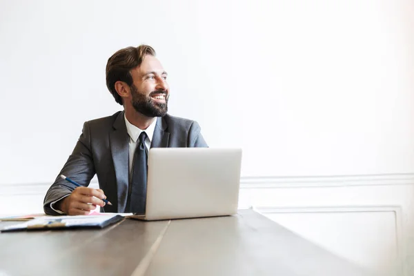 Imagem de empresário barbudo alegre trabalhando no laptop no escritório enquanto escreve notas e olha para o lado — Fotografia de Stock