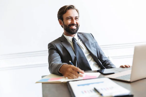 Image of smiling handsome businessman working at laptop in office while looking forward at copyspace — Stock Photo, Image