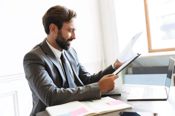 Bild eines lächelnden gutaussehenden Geschäftsmannes, der Dokumente liest, während er im Büro am Laptop arbeitet — Stockfoto