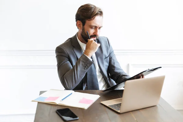 Образ мыслящего привлекательного бизнесмена, читающего документы во время работы за ноутбуком в офисе — стоковое фото