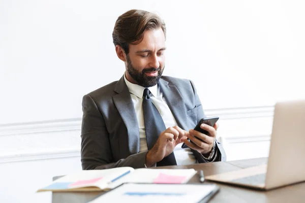 Imagem de empresário barbudo satisfeito digitando no celular enquanto trabalhava no laptop no escritório — Fotografia de Stock