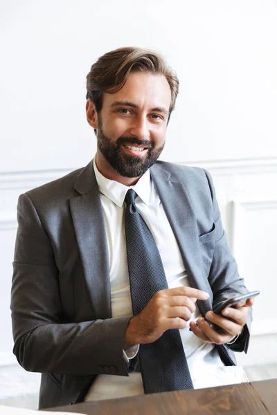 Image of smiling satisfied businessman typing on cellphone and looking at camera while working in office — Stock Photo, Image