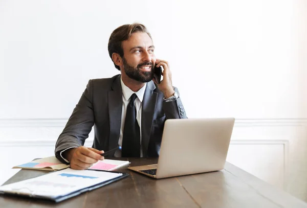 Kuva hymyilevä tyytyväinen liikemies puhuu kännykkään työskennellessään kannettavan toimistossa — kuvapankkivalokuva