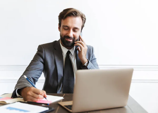 Image of smiling unshaven businessman talking on cellphone and writing down notes while working at laptop in office — Stock Photo, Image