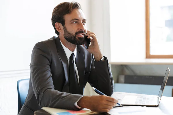 Handsome pleased optimistic bearded business man sitting in office using laptop computer working indoors talking by mobile phone. — Stock Photo, Image