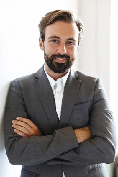 Sonriendo hombre de negocios barbudo positivo posando en la oficina cerca de la ventana en el interior . — Foto de Stock