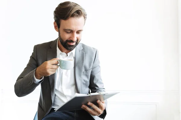 Handsome optimistic smiling bearded business man in office drinking coffee holding clipboard. — Stock Photo, Image