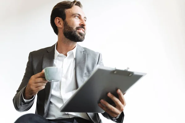 Beau homme d'affaires barbu optimiste dans le bureau boire du café tenant presse-papiers . — Photo