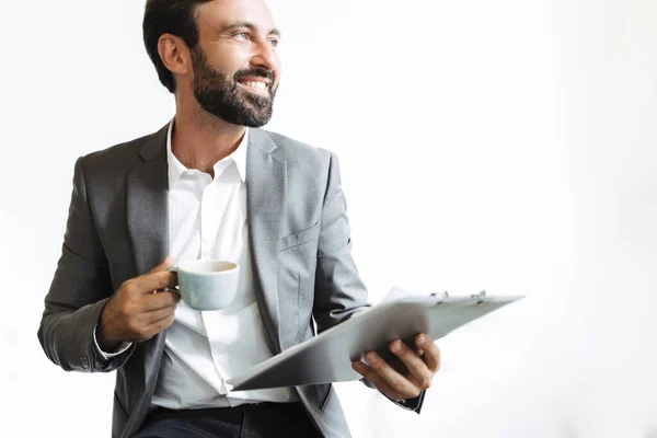 Image of smiling satisfied businessman drinking coffee and holding clipboard while working in office — 图库照片