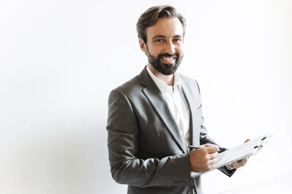 Image of smiling successful businessman holding clipboard and writing down notes while working in office — Stock Photo, Image