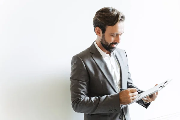 Image of satisfied successful businessman holding clipboard and writing down notes while working in office — ストック写真